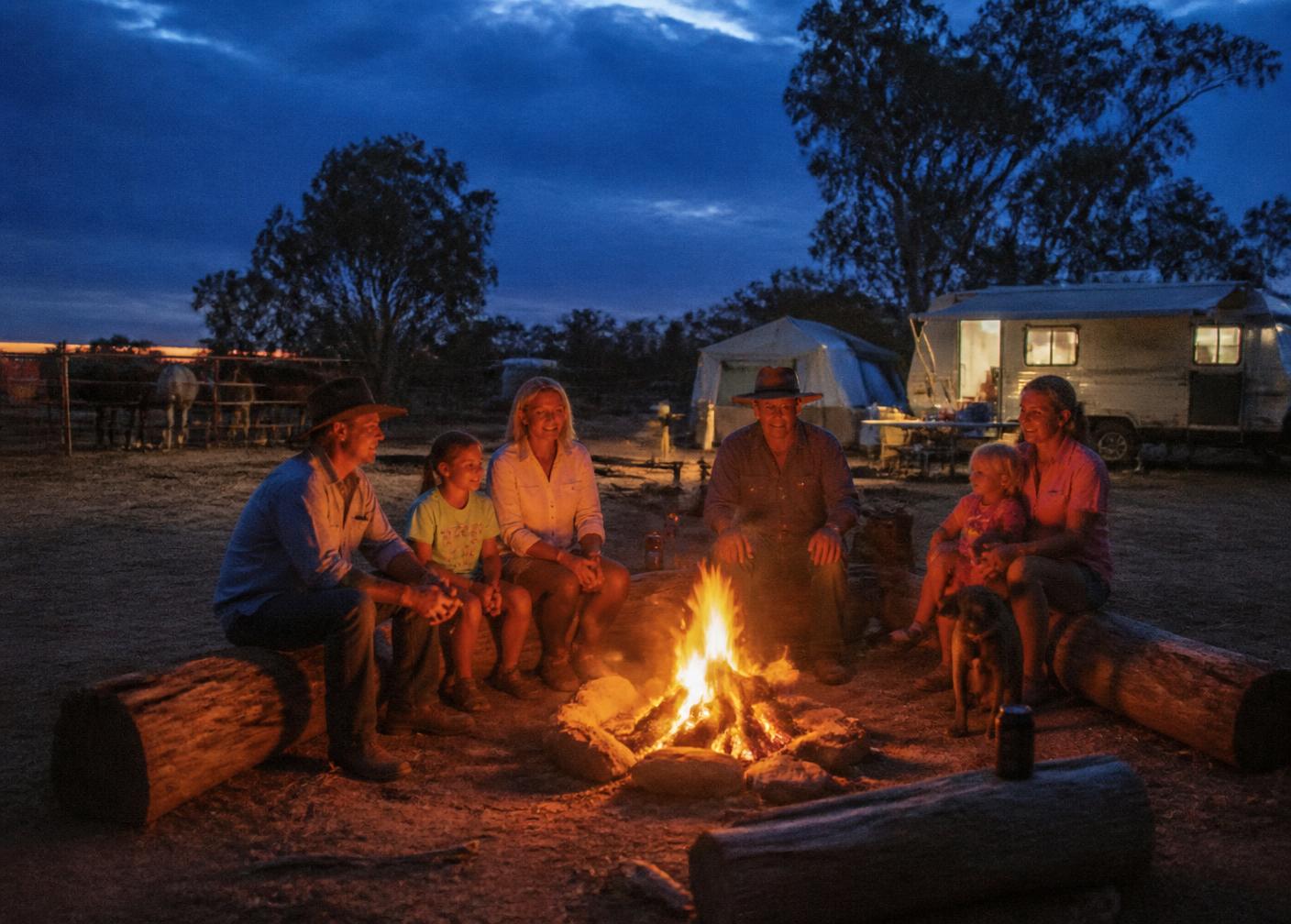Families sitting around the campfire at dusk with tents, a caravan and horse yards behind